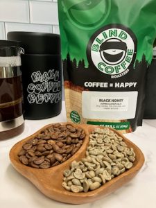 Photo showing close up of Black Honey coffee beans green and roasted with bag of Black Honey, French press, coffee canister, and small coffee tree in the background.