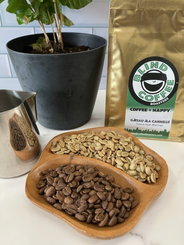 Photo showing close up of green and roasted Gateau a la Cannelle beans with bag of Gateau a la Cannelle, milk pitcher, and small coffee tree in the background.